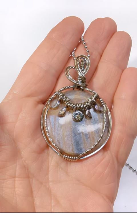Black moonstone circular gemstones pendant with mesmerising blueish silver flash, set in a unique sterling silver twisted wire wrap setting and hanging from a stainless steel ball chain included for free in any size. This elegant gemstone comes in orange, black, browns, rust , greys and silver tones all mixed together like a dance of colours with a magnificent silver and blue flash from all directions when looked upon.